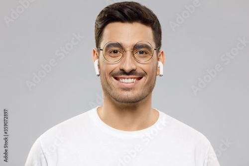 Close-up portrait of young handsome smiling man wearing white t-shirt and wireless earphones, enjoying favorite music, isolated on gray background