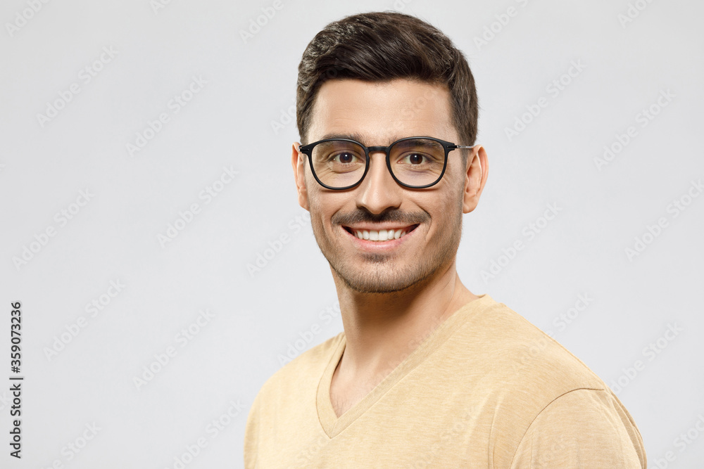 Portrait of young handsome smiling man dressed in beige t-shirt, wearing glasses, isolated on gray background Portrait of young handsome smiling man dressed in beige t-shirt, wearing glasses, isolated on gray background