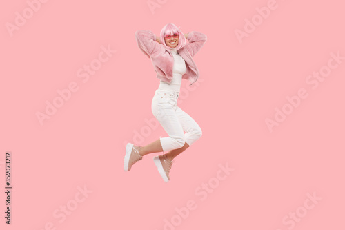 Full length portrait of young active woman wearing colored wig and glasses, jumping in air while having fun at party, isolated on pink background