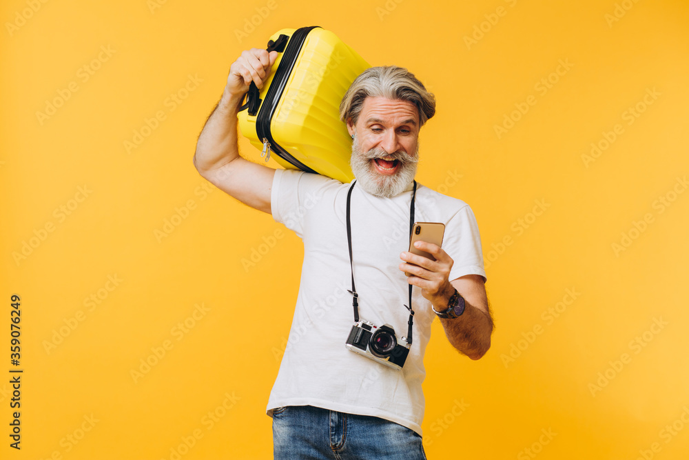 Stylish bearded man laughing, holding a yellow suitcase and looking at the phone choosing a country to travel.