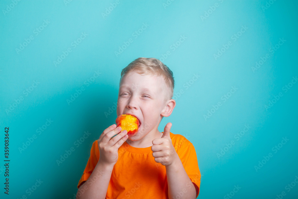 Cute and funny baby boy in an orange t-shirt bites off an orange juicy peach. The child is a blond European on a blue background. Health benefits of fresh fruit. Cute and funny baby boy in an orange t-shirt bites off an orange juicy peach. The child is a blond European on a blue background. Health benefits of fresh fruit.