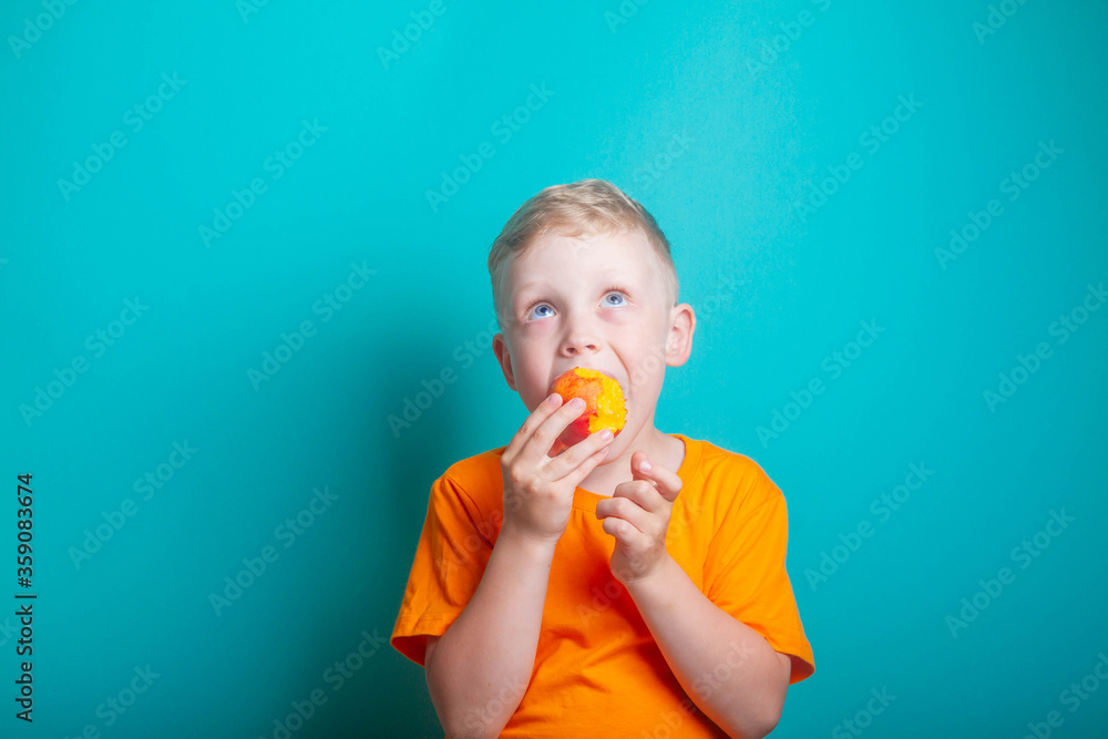 Cute and funny baby boy in an orange t-shirt bites off an orange juicy peach. The child is a blond European on a blue background. Health benefits of fresh fruit. Cute and funny baby boy in an orange t-shirt bites off an orange juicy peach. The child is a blond European on a blue background. Health benefits of fresh fruit.