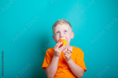 Cute and funny baby boy in an orange t-shirt bites off an orange juicy peach. The child is a blond European on a blue background. Health benefits of fresh fruit.