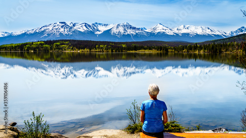Senior woman sitting at Pyramid Lake with Mount Edith Cavell, Aquila Mountain, Majestic Mountain and other snow capped peaks in the background in Jasper National Park in Alberta, Canada