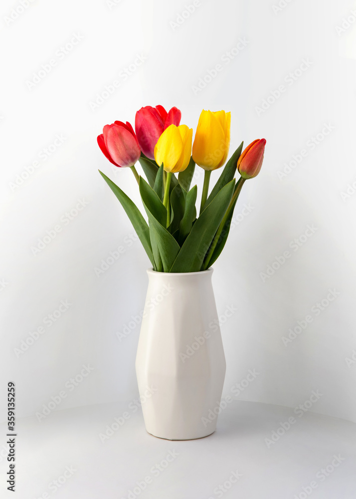Red and yellow tulips in a white geometric ceramic vase stand. Flowers in a vase. mother's day. Bouquet of flowers in the morning sun. Yellow and red tulips stand in a white vase on a gray background Red and yellow tulips in a white geometric ceramic vase stand. Flowers in a vase. mother's day. Bouquet of flowers in the morning sun. Yellow and red tulips stand in a white vase on a gray background