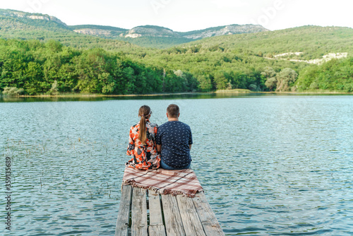 The couple sits on a wooden pier on the lake. There is a green forest and mountains around the lake. A guy in a blue T-shirt, a girl in a motley dress, sitting on a plaid. The concept of rest, travel.