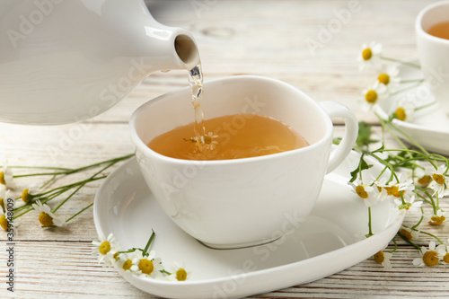 Pouring tasty chamomile tea into cup on white wooden table