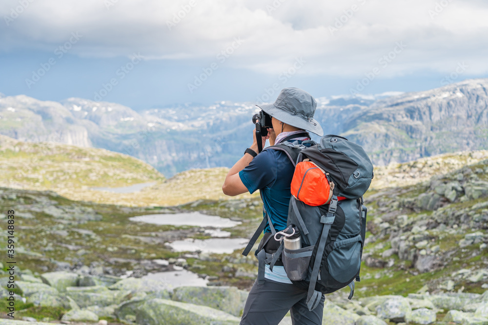 Young Asian man traveller with backpack taking picture of mountains landscape while trekking in Trolltunga mountain cliff, famous trekking route in Odda, Norway, Scandinavia