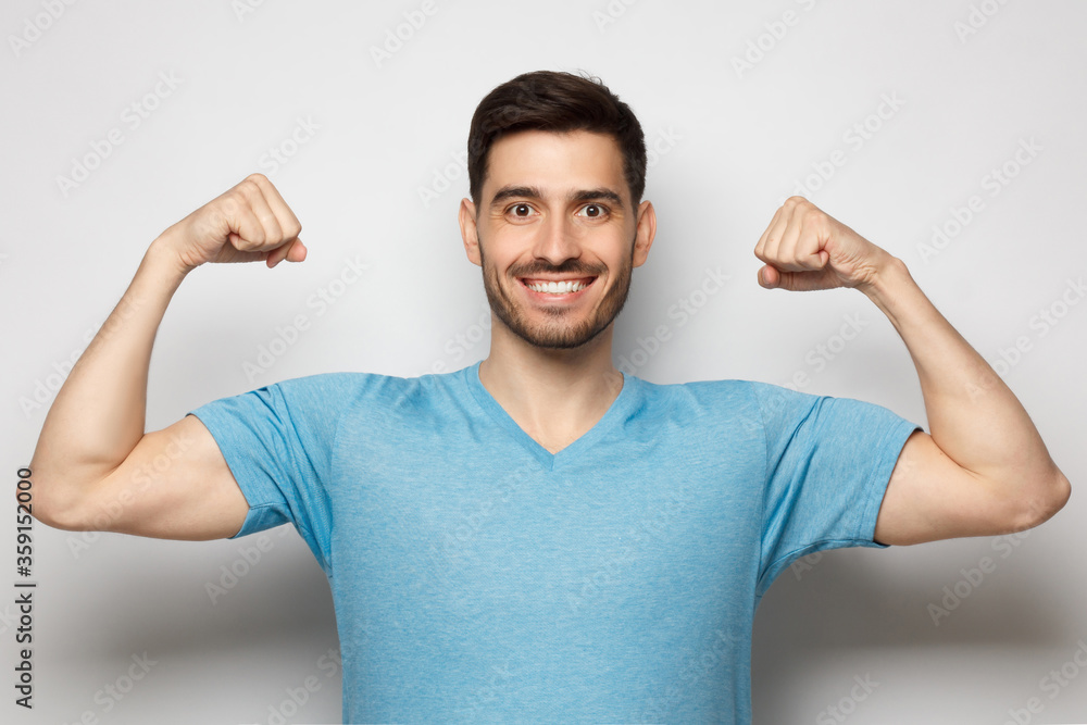 Strong and sporty athletic man in casual blue t-shirt, showing two biceps after training in gym, isolated on gray background Strong and sporty athletic man in casual blue t-shirt, showing two biceps after training in gym, isolated on gray background