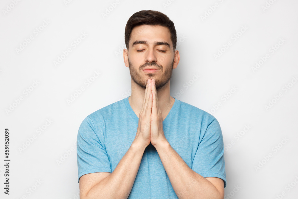 Close up of young man putting hands together as if he is praying with closed eyes, isolated on gray background Close up of young man putting hands together as if he is praying with closed eyes, isolated on gray background