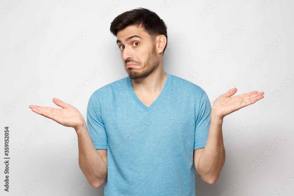 I don't know. Portrait of young confused man in blue t-shirt standing and shrugging shoulders, spreading hands isolated on gray background I don't know. Portrait of young confused man in blue t-shirt standing and shrugging shoulders, spreading hands isolated on gray background