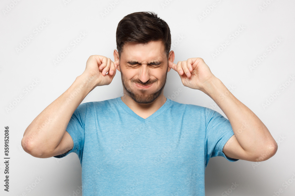 Young man in blue tshirt closing ears suffering from loud noise, isolated on gray background Young man in blue tshirt closing ears suffering from loud noise, isolated on gray background
