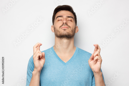 Young man with eyes closed and crossed fingers of both hands, wishing luck, isolated on gray background