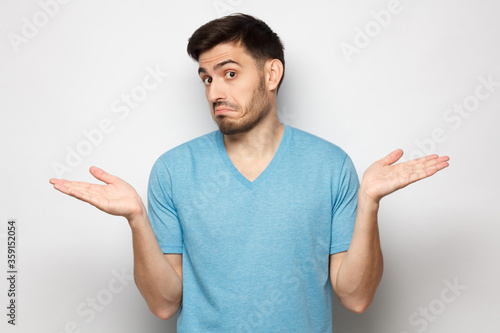 I don't know. Portrait of young confused man in blue t-shirt standing and shrugging shoulders, spreading hands isolated on gray background photo