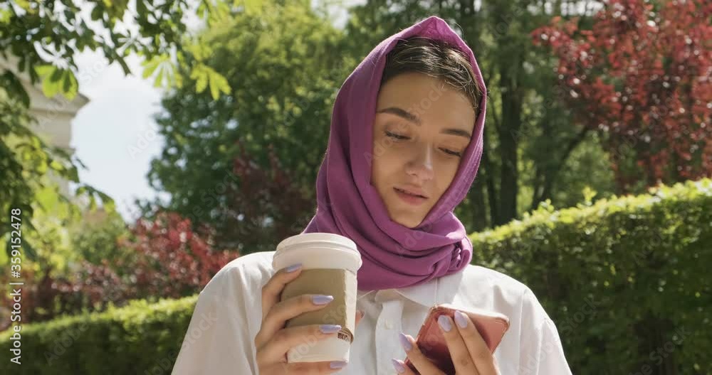 Beautiful young woman drink coffee and looking at smartphone, wearing traditional headscarf. Attractive Female in hijab Beautiful young woman drink coffee and looking at smartphone, wearing traditional headscarf. Attractive Female in hijab