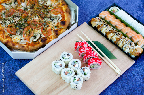 A lot of different sushi rolls and a big pizza in a box. Sushi rolls, green leaf and sticks. photo