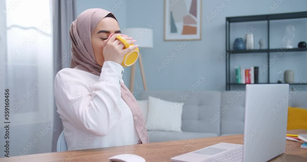 Middle plan of young muslim woman using laptop computer at home. Portrait of business woman in hijab watching news at laptop screen and drinking coffee and smiling to camera at home workplace. Middle plan of young muslim woman using laptop computer at home. Portrait of business woman in hijab watching news at laptop screen and drinking coffee and smiling to camera at home workplace.
