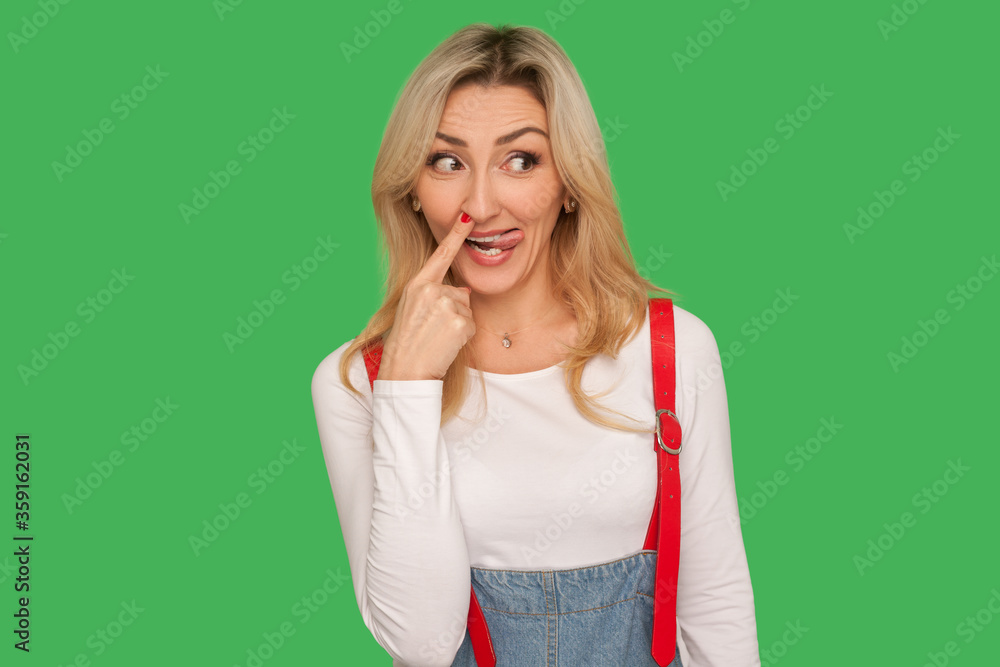 Portrait of crazy funny adult woman in denim overalls picking out boogers, drilling nose and smiling with comical humorous expression, fooling around. indoor studio shot isolated on green background Portrait of crazy funny adult woman in denim overalls picking out boogers, drilling nose and smiling with comical humorous expression, fooling around. indoor studio shot isolated on green background