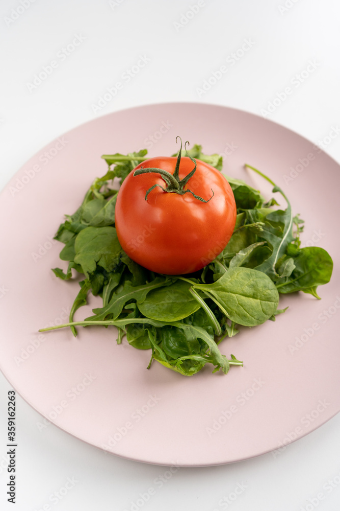 Salad cooked with fresh spinach and arugula leaves and tomato on a pink plate in the center on a white table. Healthy diet. Close-up. Vertical Salad cooked with fresh spinach and arugula leaves and tomato on a pink plate in the center on a white table. Healthy diet. Close-up. Vertical