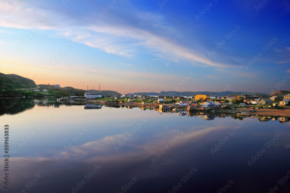 Summer evening in Teriberka, Murmansk oblast, Kola peninsula, Northern Russia. Beautiful nature scenery, sunset sky with cloud reflected in smooth surface of calm water, surf, wooden houses and boats