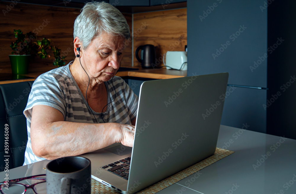 An elderly woman in headphones sits at the table and prints on a laptop in the home environment. Concept of silver generation and gadgets. Seniors and technology. An elderly woman in headphones sits at the table and prints on a laptop in the home environment. Concept of silver generation and gadgets. Seniors and technology.