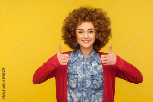 Like gesture! Happy successful woman with curly hair in casual style jeans shirt showing thumbs up, satisfied with excellent result, good feedback. indoor studio shot isolated on yellow background