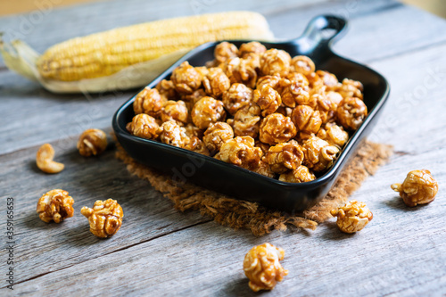 Flat lay of tasty caramel popcorn in black ceramic pan plate and corn on wooden table, close up.