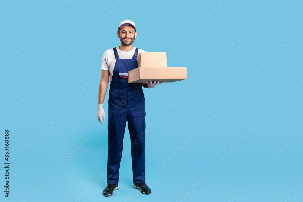 Full length delivery man in blue uniform with gloves standing, holding cardboard boxes and smiling to camera. Profession of courier, shipment and cargo transportation service. studio shot isolated Full length delivery man in blue uniform with gloves standing, holding cardboard boxes and smiling to camera. Profession of courier, shipment and cargo transportation service. studio shot isolated