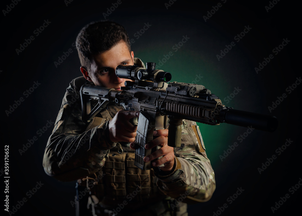 a brutal guy in military airsoft overalls poses with a weapon in his hands on a dark background