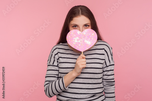 Be my Valentine! Portrait of coquettish cheerful woman peeping out shiny pink heart with playful happy look, demonstrating love care, romantic feelings. indoor studio shot isolated on pink background