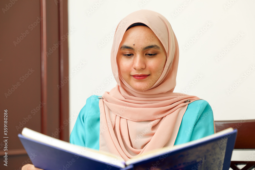 young Asian Malay Muslim woman wearing headscarf at home office student sitting at table read book document note study smile happy look