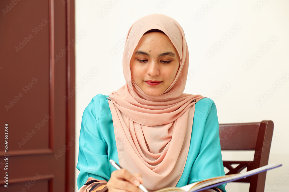 young Asian Malay Muslim woman wearing headscarf at home office student sitting at table read write book document note study smile happy look young Asian Malay Muslim woman wearing headscarf at home office student sitting at table read write book document note study smile happy look