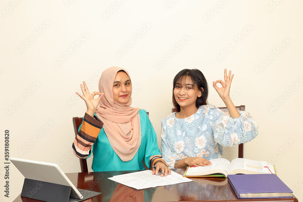 Two young Asian Malay Muslim woman wearing headscarf at home office student sitting at table talk mingle computer book document study ok finger sign look at camera Two young Asian Malay Muslim woman wearing headscarf at home office student sitting at table talk mingle computer book document study ok finger sign look at camera