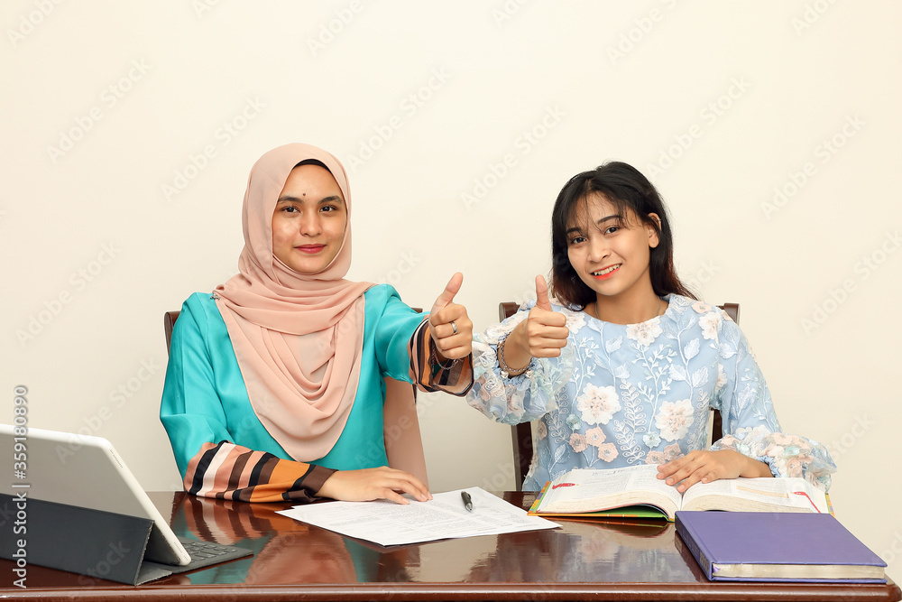 Two young Asian Malay Muslim woman wearing headscarf at home office student sitting at table talk mingle computer book document study thumbs up sign look at camera Two young Asian Malay Muslim woman wearing headscarf at home office student sitting at table talk mingle computer book document study thumbs up sign look at camera