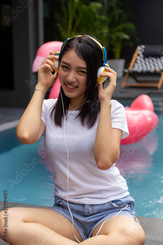 Young woman using headphone for listening nice music with swimming pool of resort or hotel background with happiness, smile face. Asian pretty girl love listen music that make her relieve and relax