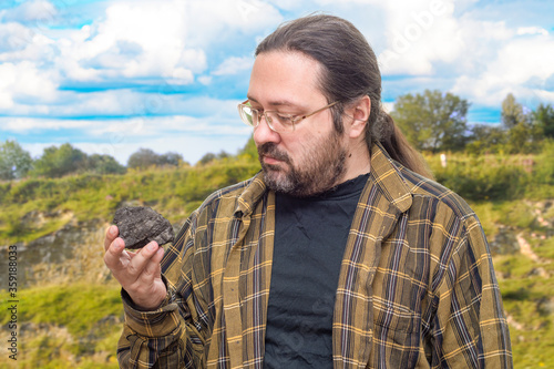 A geologist man in clean clothes with glasses holds in his hand a piece of coal on a blurry background of mountainous terrain. Illustration of research on fossil fuels with a non-renewable resource. © Николай Батаев