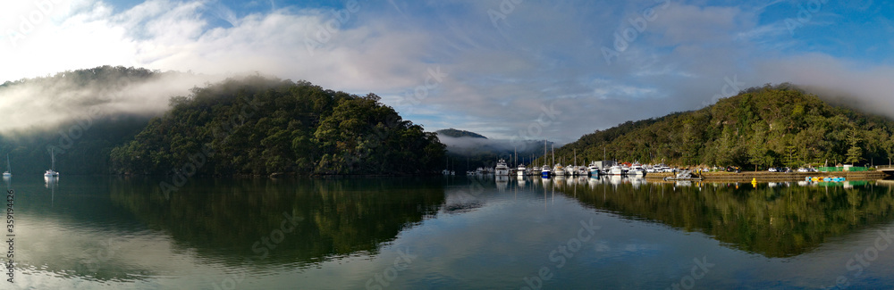 Beautiful morning panoramic view of Cockle creek with reflections of blue sky, light clouds, foggy mountains and trees, Bobbin Head, Ku-ring-gai Chase National Park, New South Wales, Australia