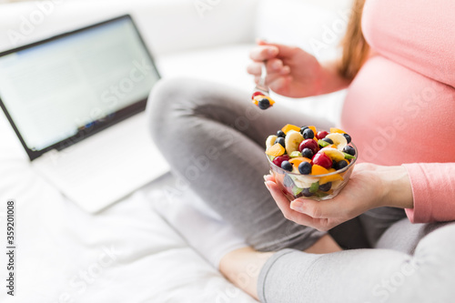 Close up of pregnant woman eating various fruits salad while working on laptop.