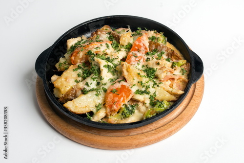 Dish on a black pan, which stands on a wooden stand. Potatoes with cheese and red pepper. Sprinkled with green parsley.