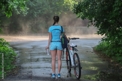 sport girl in sportswear stands with her back in front of a bicycle in her hand a water bottle and looks forward