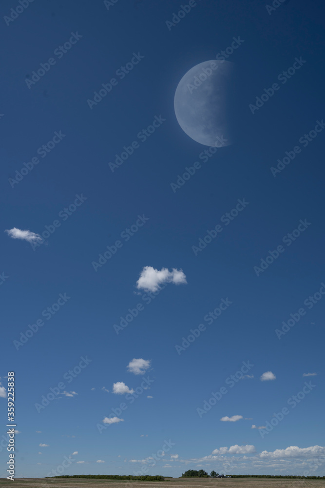 Dramatic photograph of a rural township on the Alberta Prairies just after sunrise with big white clouds on a blue sky background with a daytime partial moon in a rcommunity. Dramatic photograph of a rural township on the Alberta Prairies just after sunrise with big white clouds on a blue sky background with a daytime partial moon in a rcommunity.