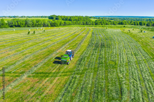 An agricultural tractor collects mowed grass for agricultural use and wraps hay bales in a plastic field  rural landscape aerial view.