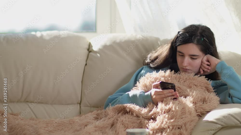 Cute brunette girl sitting on the couch during the day and reading the news on her phone, copy space Cute brunette girl sitting on the couch during the day and reading the news on her phone, copy space