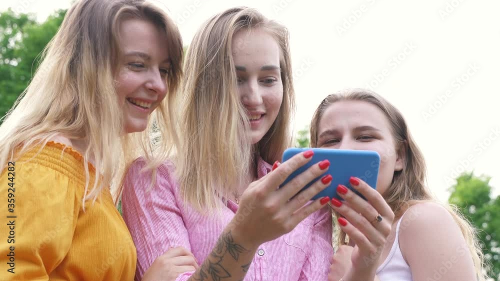 Portrait of Three Beautiful Girls Look At Phone, Laugh. Young Womans Watch Social Media Video In Mobile Smart Phone. Girlfriends Looking at Smartphone. Summer Outdoors Countryside Park Friends Party. Portrait of Three Beautiful Girls Look At Phone, Laugh. Young Womans Watch Social Media Video In Mobile Smart Phone. Girlfriends Looking at Smartphone. Summer Outdoors Countryside Park Friends Party.