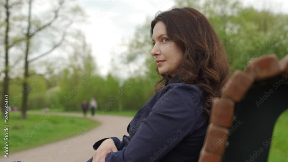 Beautiful adult brunette woman sitting on bench in a park, portrait of woman with long hair on summer day. Beautiful adult brunette woman sitting on bench in a park, portrait of woman with long hair on summer day.