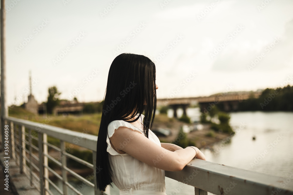 A girl in a white dress walks on the street. A girl stands on the bridge and looks into the distance. The woman has black hair.
