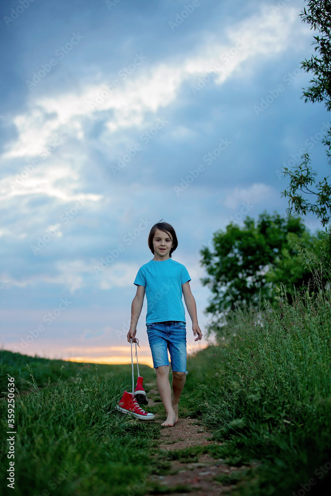 Happy child, holding pair of sneakers in hands, walking on a rural path