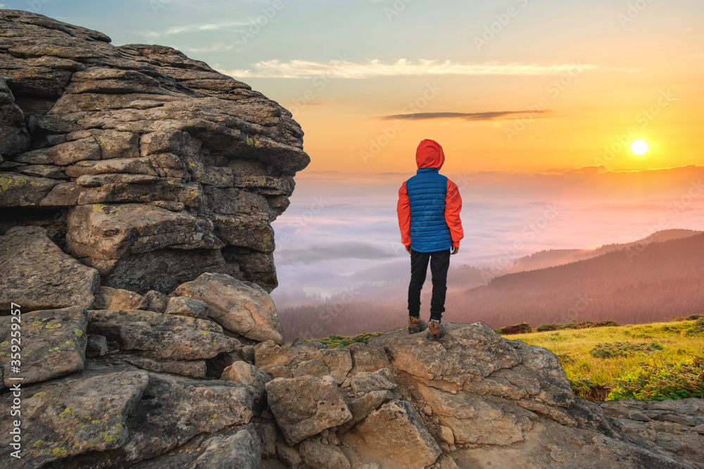 Young child boy hiker standing in mountains enjoying view of amazing mountain landscape at sunrise.