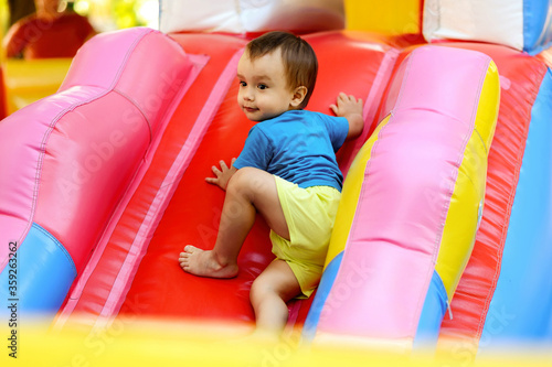 Fotografie Toddler boy in blue shirt and yellow shorts climbing up slide on bright inflatable bouncy castle
