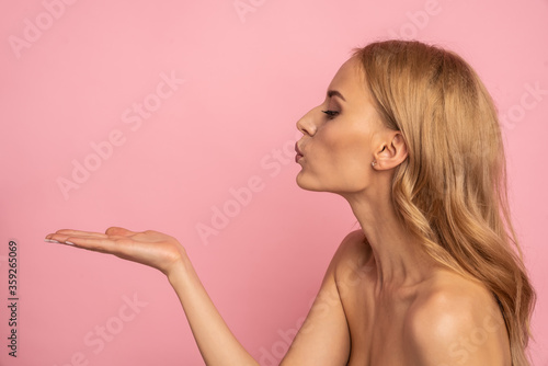 Profile portrait of cute, charming model blowing kiss with pout lips holding copyspace empty place on one palm looking at product isolated on pink background, advertisement concept